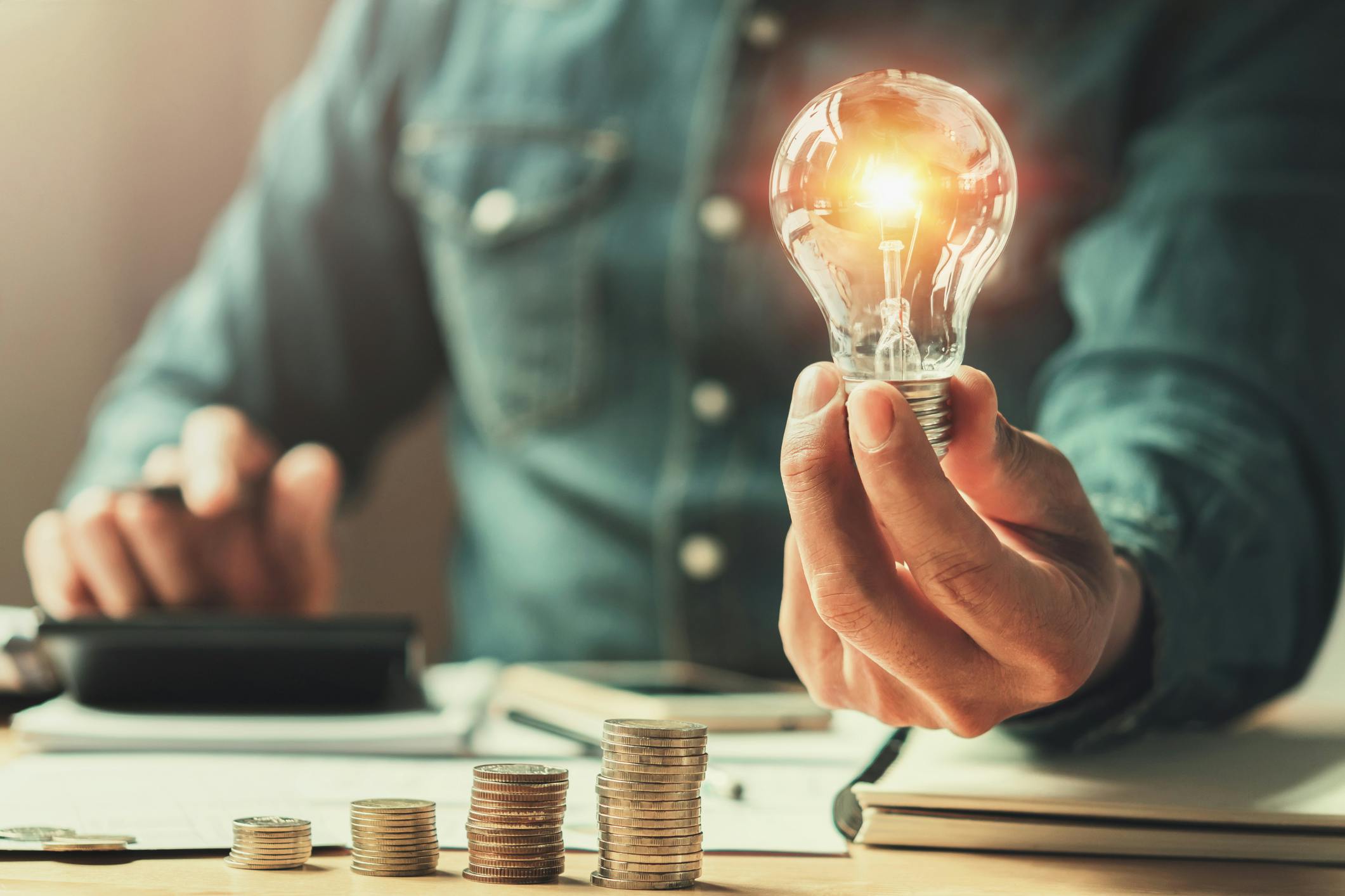 Man holds a light bulb in one hand while doing calculations behind a stack of coins