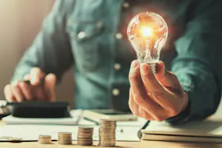 Man holds a light bulb in one hand while doing calculations behind a stack of coins