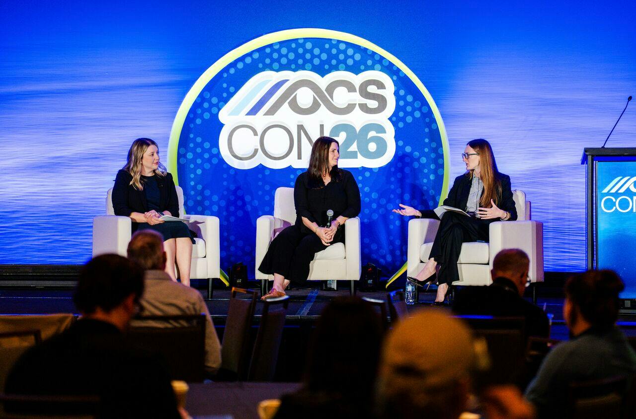 Three women speak on a stage in front of an OCS CON logo.
